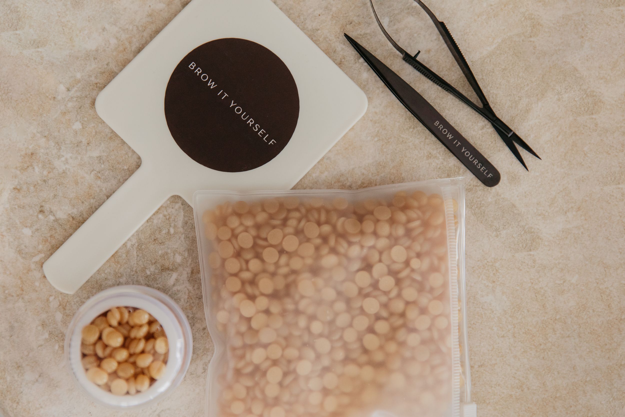 Brew it yourself kit with wax beads in bag, small bowl of beads, tweezers, and mirror on marble surface.