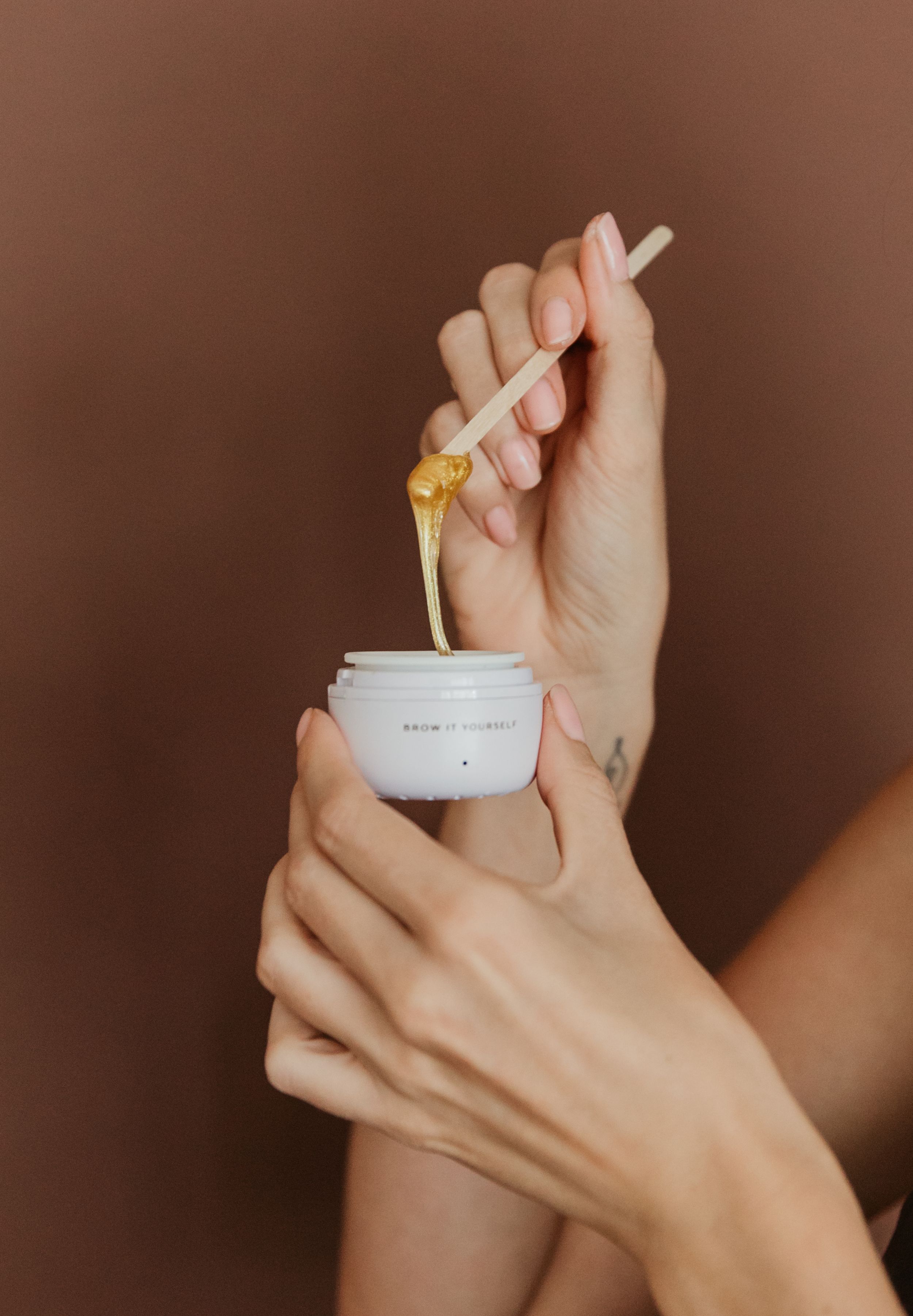 Hands holding a white jar of honey balm with a wooden stick dripping golden honey on a brown background.