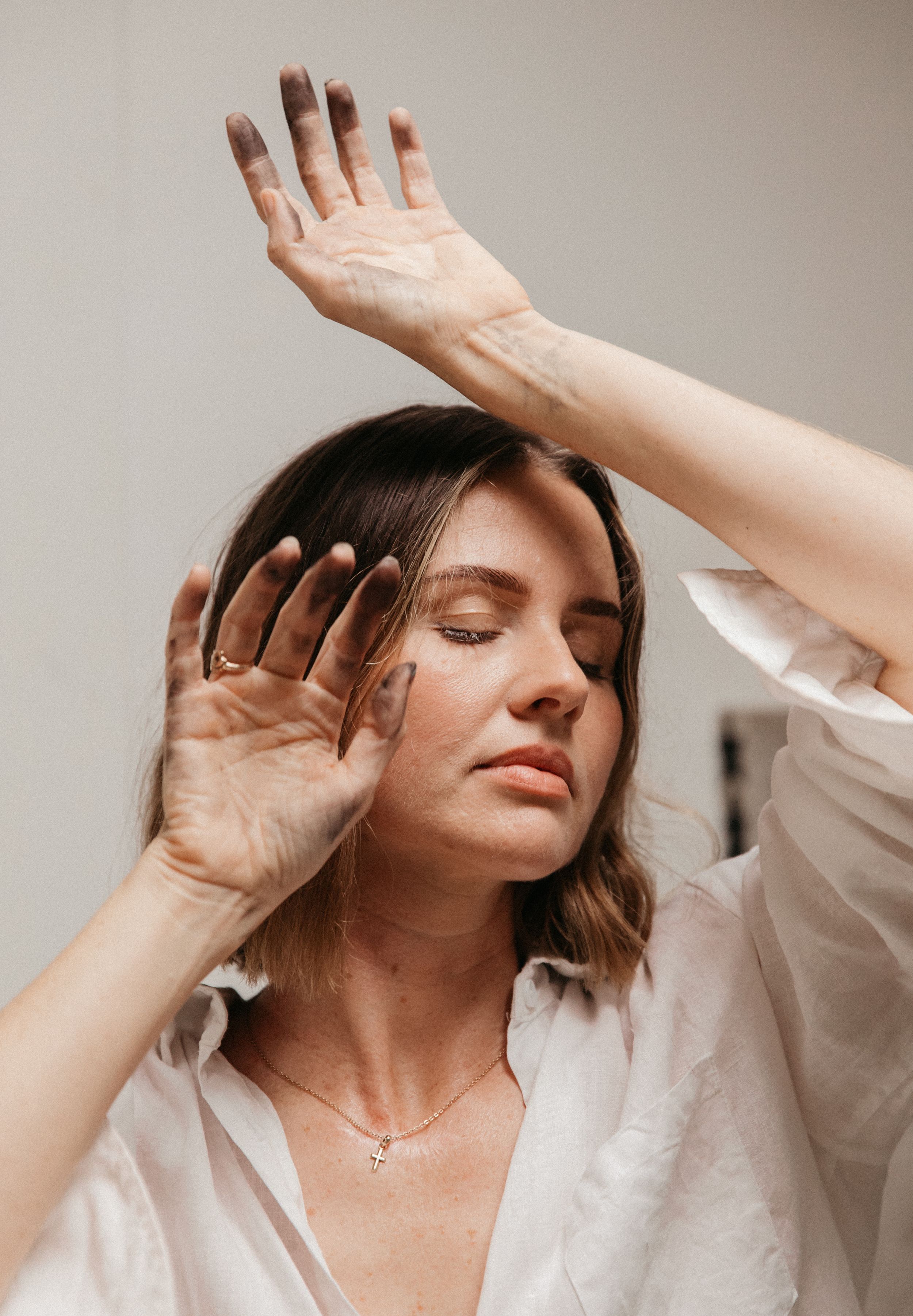 Woman in white shirt with eyes closed, hands raised near face, wearing a cross necklace, in a serene pose.