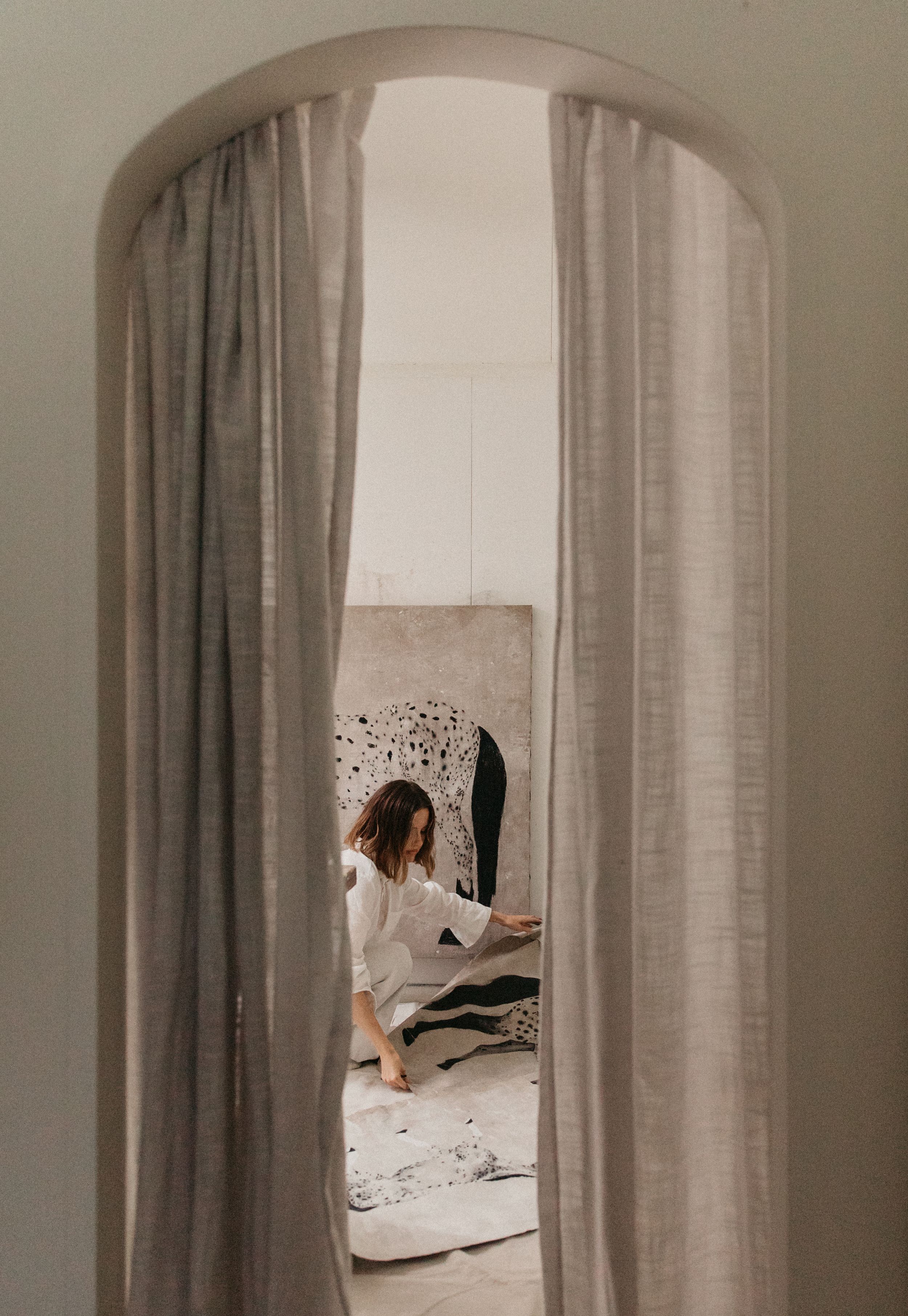 Woman in white outfit sitting on floor viewed through curved curtained archway in minimalist interior.