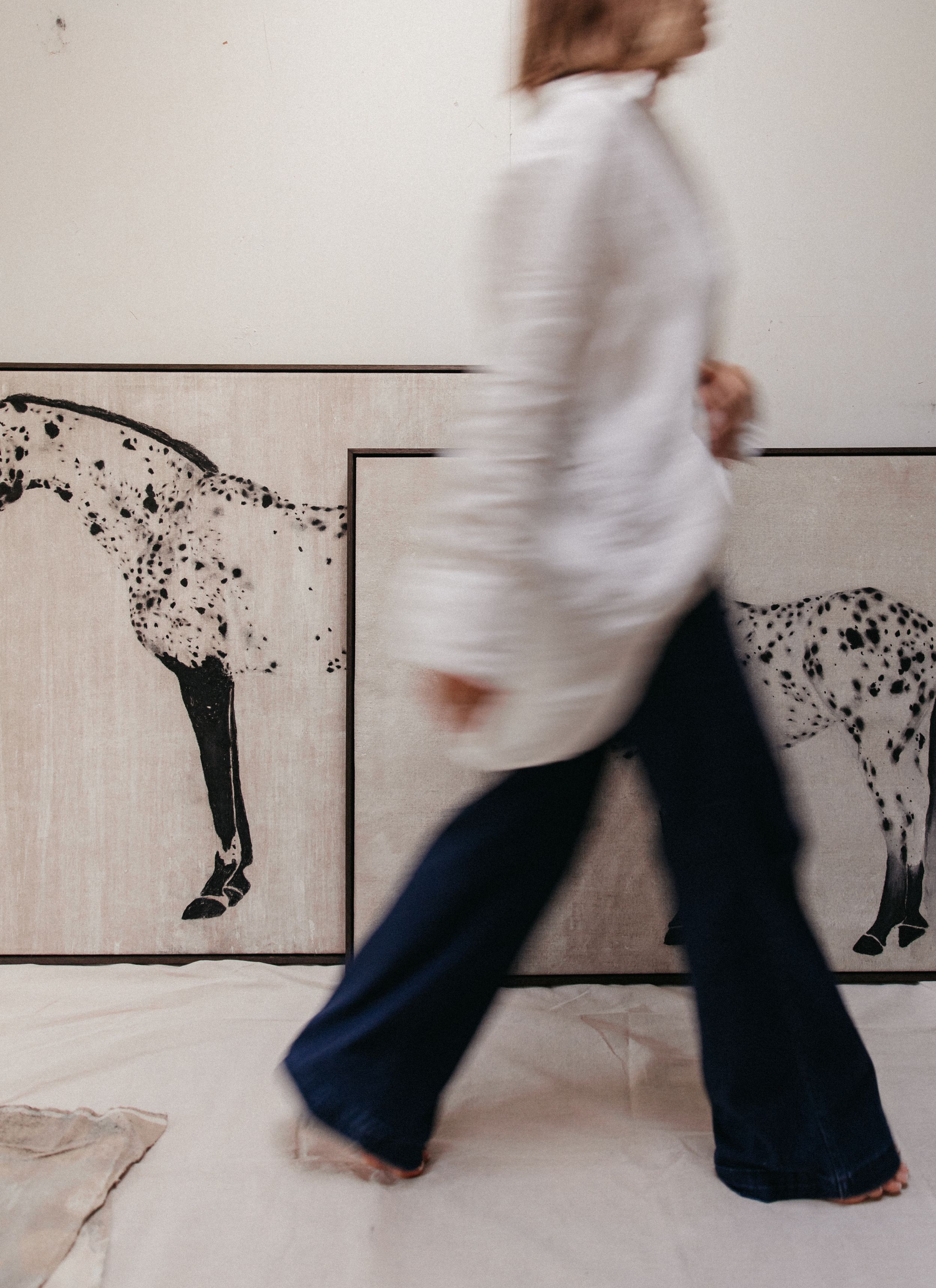 Blurred woman in white shirt and flare jeans walking past two large horse paintings in an art studio.
