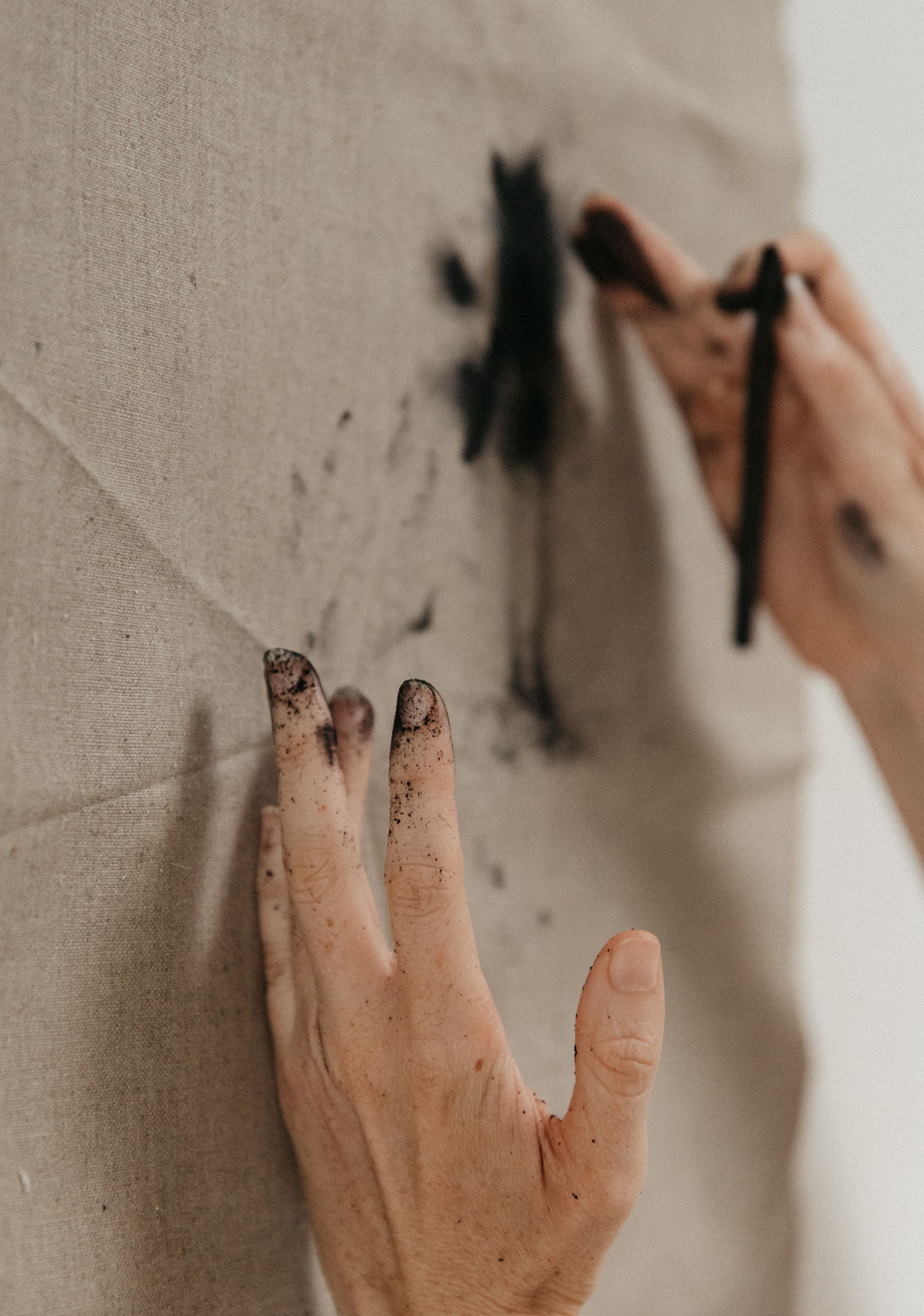 Artist's hands with black paint-stained fingers creating abstract brushstrokes on natural linen canvas.