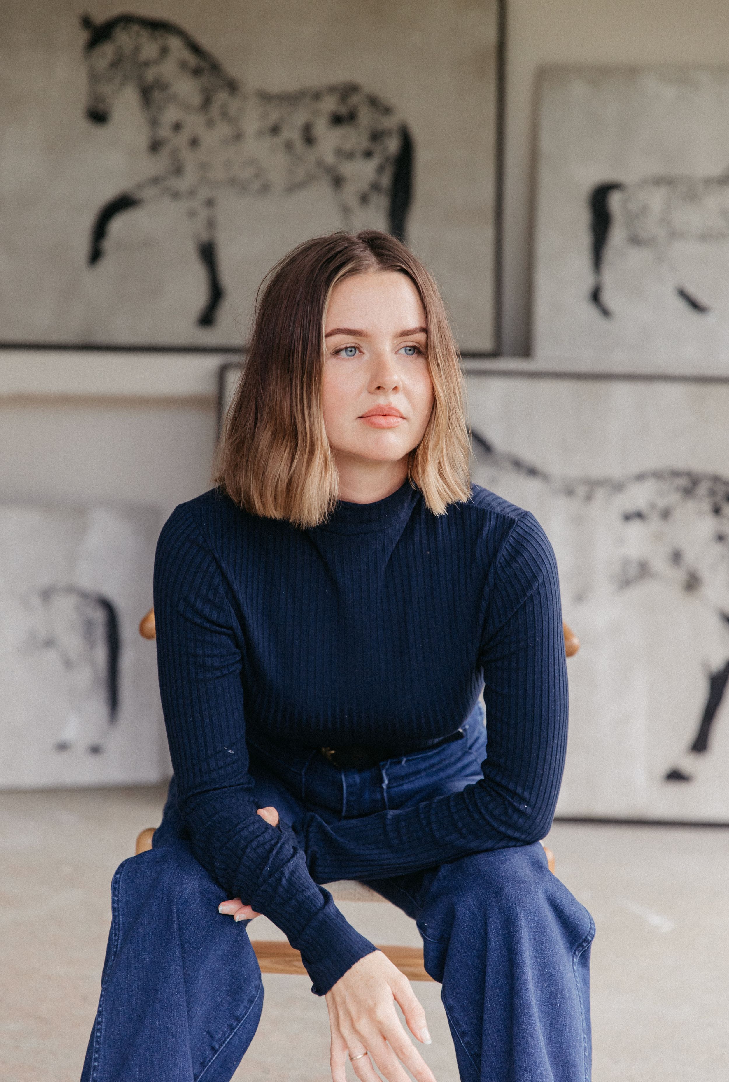 Young woman with bob haircut in navy ribbed turtleneck and jeans sits on stool before horse paintings.