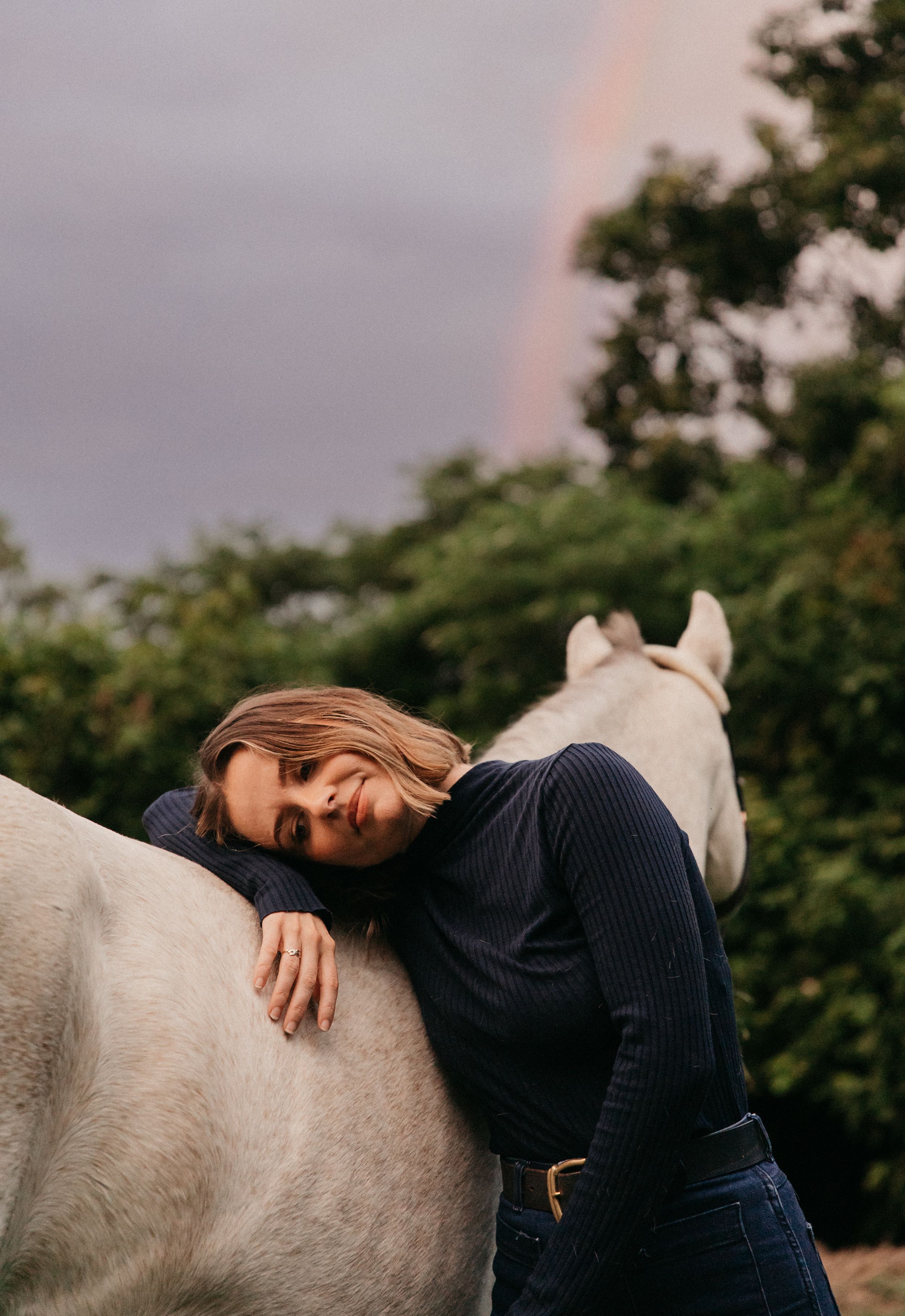 Woman leaning on a white horse with a rainbow in the background among green trees.
