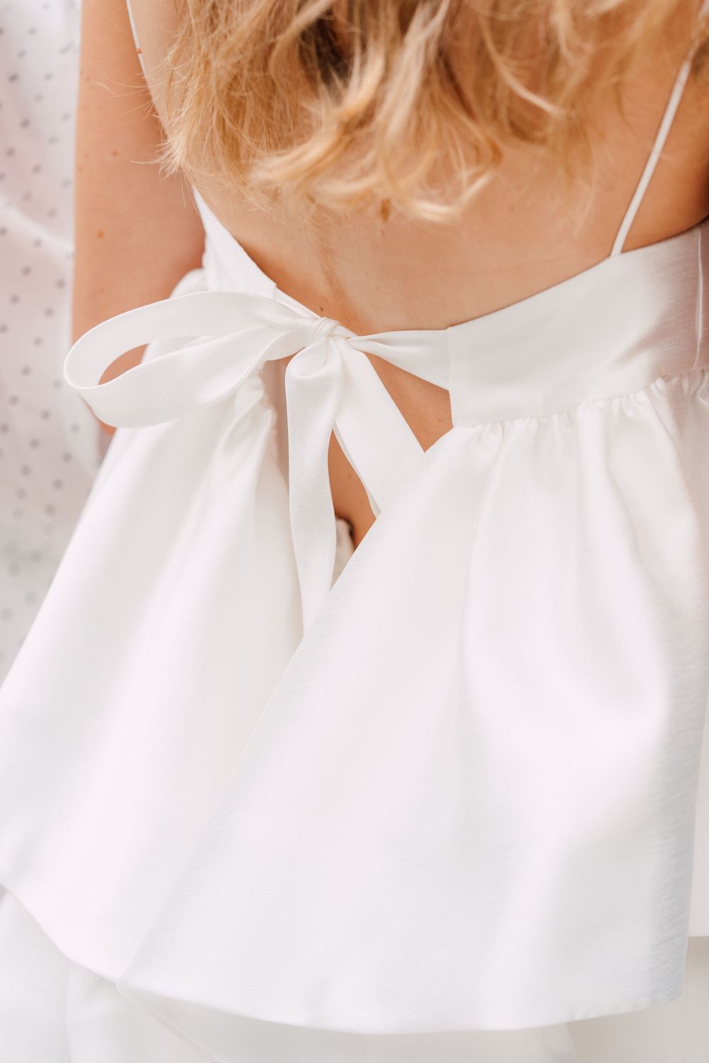 White satin camisole top with elegant bow tie back detail and spaghetti straps, shown on a blonde model.
