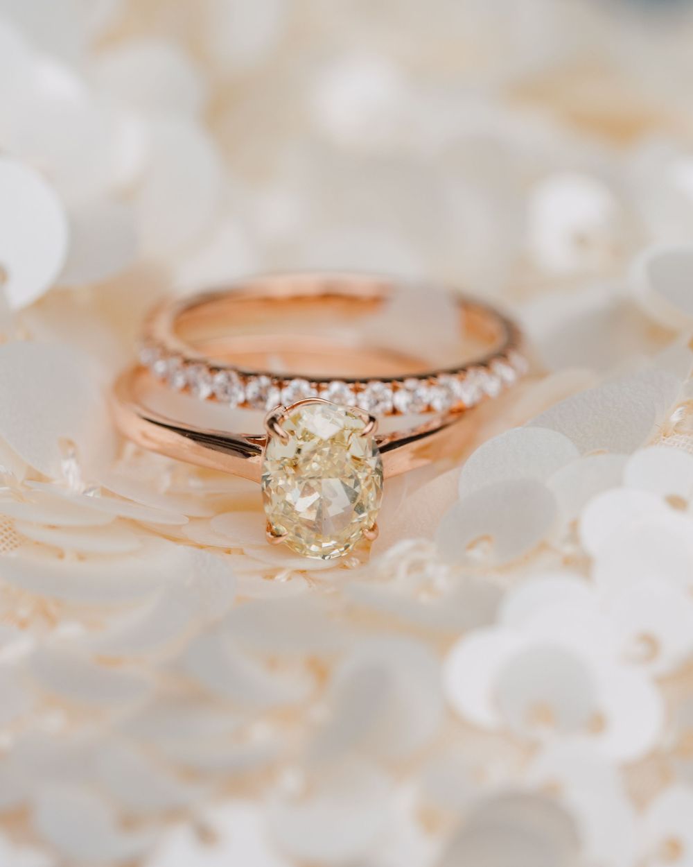Rose gold engagement ring with oval yellow diamond solitaire and diamond-studded band on white floral background.