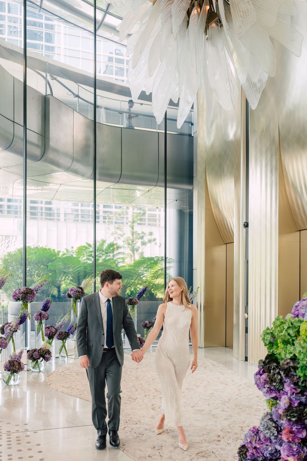 Elegant couple holding hands in a luxurious hotel lobby adorned with purple floral arrangements and a feather chandelier.