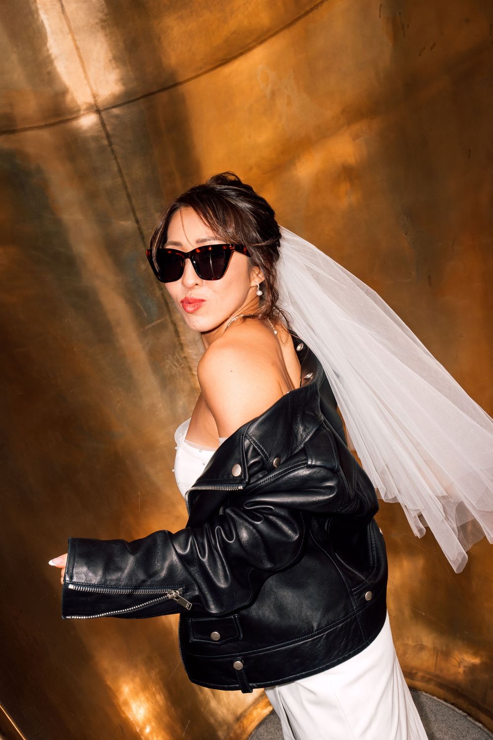 A bride wearing a white dress poses in sunglasses and a black leather jacket with a flowing veil against a warm copper background.