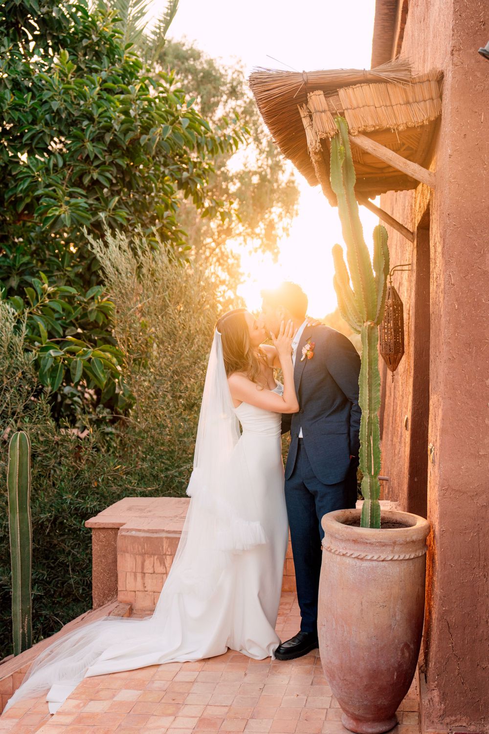 Romantic sunset moment between a couple in wedding attire by a terracotta wall and potted cactus.