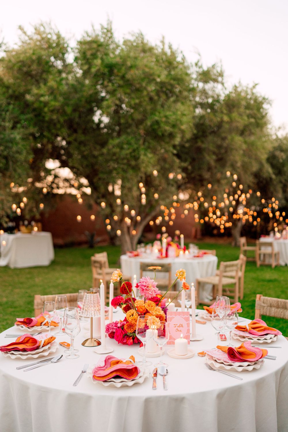 Elegant outdoor wedding reception table with pink and orange floral centerpieces under string lights.