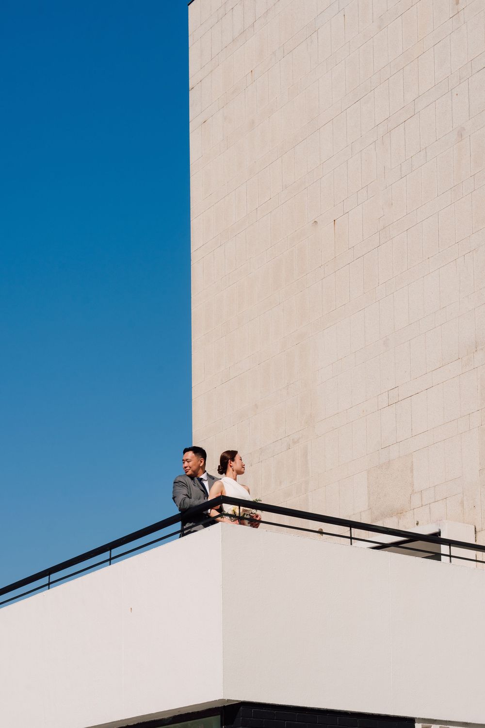 Couple standing on a modern white building balcony against a clear blue sky. Hong Kong city hall wedding photography
