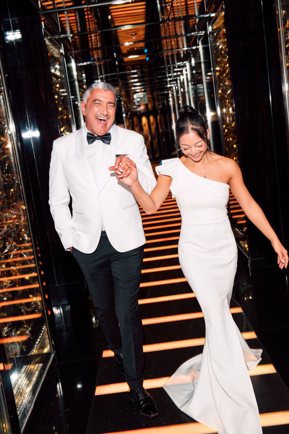Bride and groom descending mirrored hallway while laughing together.