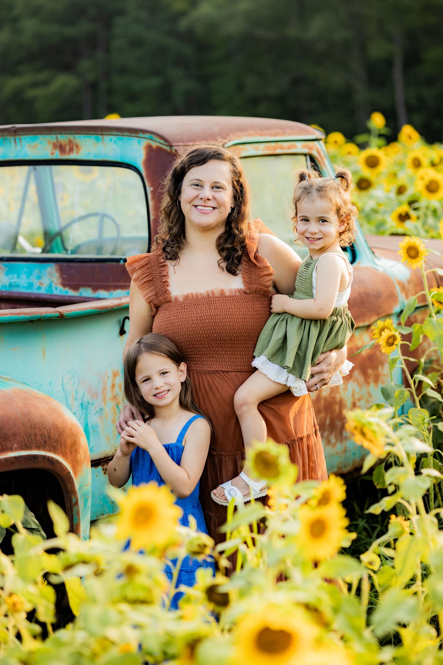 Woman and two girls posing in a sunflower field beside a vintage rusty turquoise truck on a sunny day.