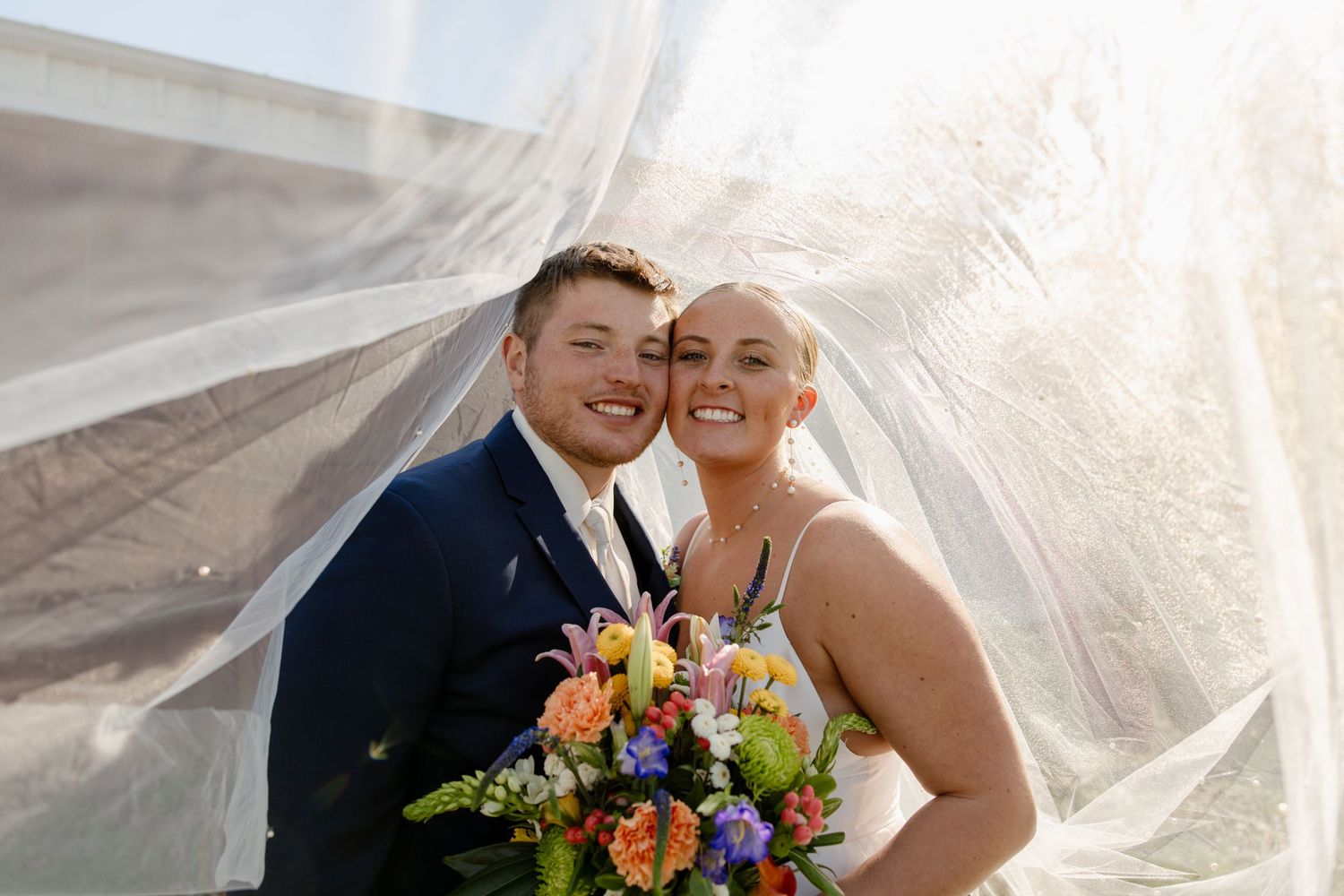 A couple shares a joyful moment under a flowing veil with colorful wedding flowers.