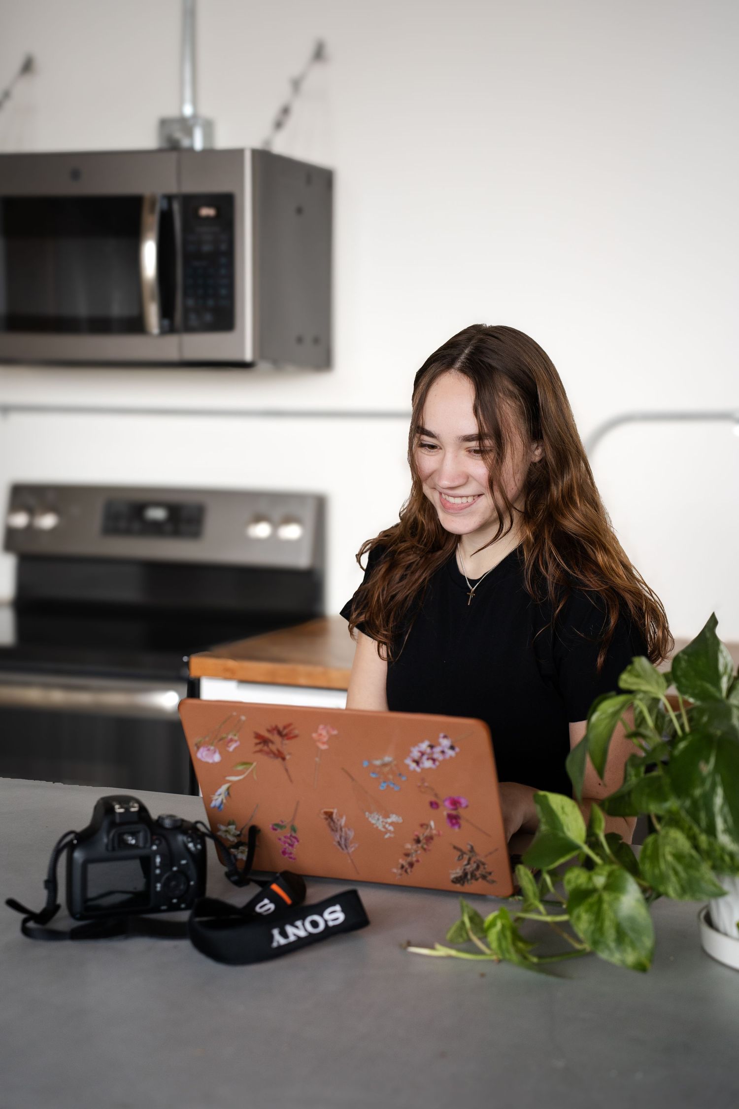 Person working on decorated laptop with camera equipment in bright modern kitchen.