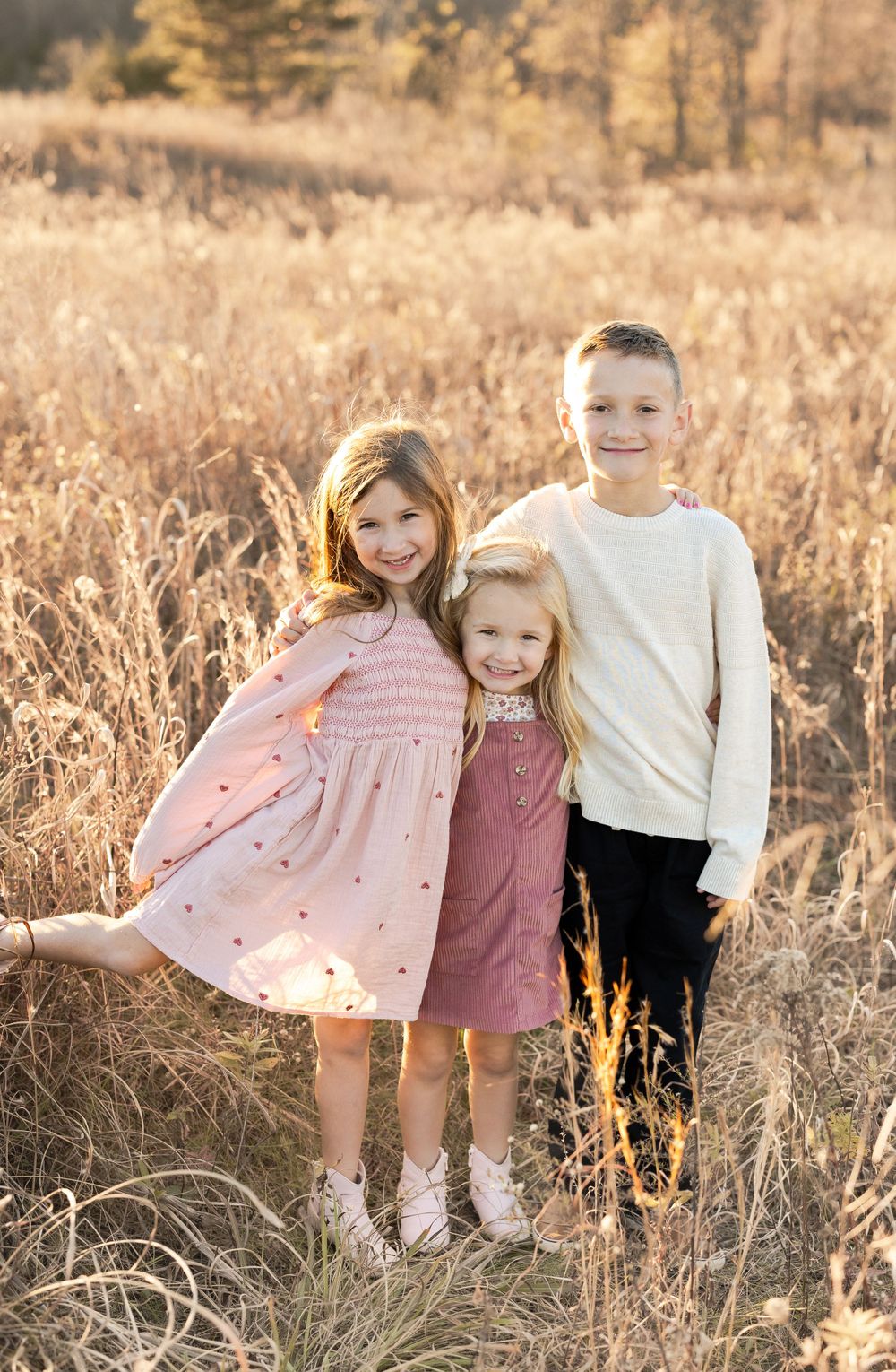 Three children standing together in tall golden grass during sunset.