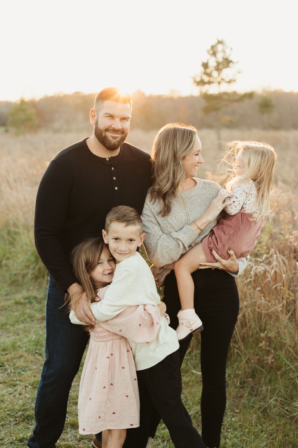 Family of five embraces during golden hour sunset in a field of tall grass.