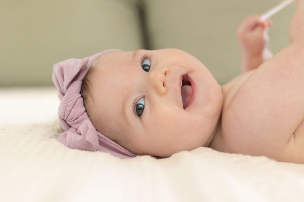 Baby wearing purple headband lying on white bedding with bright eyes and open mouth.