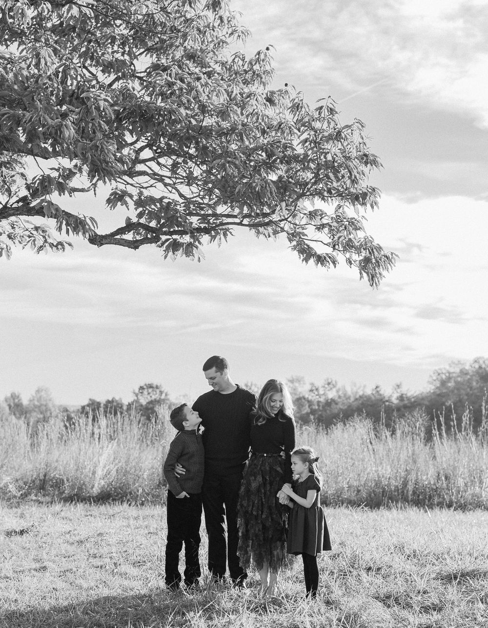 A group of people stand together in a grassy field under a large tree branch on an overcast day.