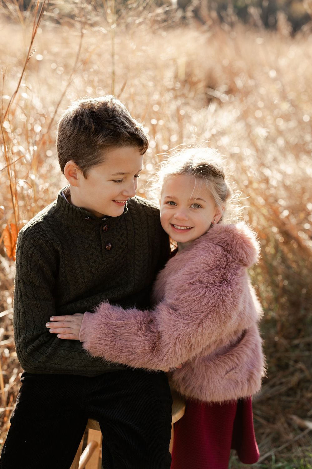 Two children in winter coats share a sweet embrace in a golden sunlit field during autumn.