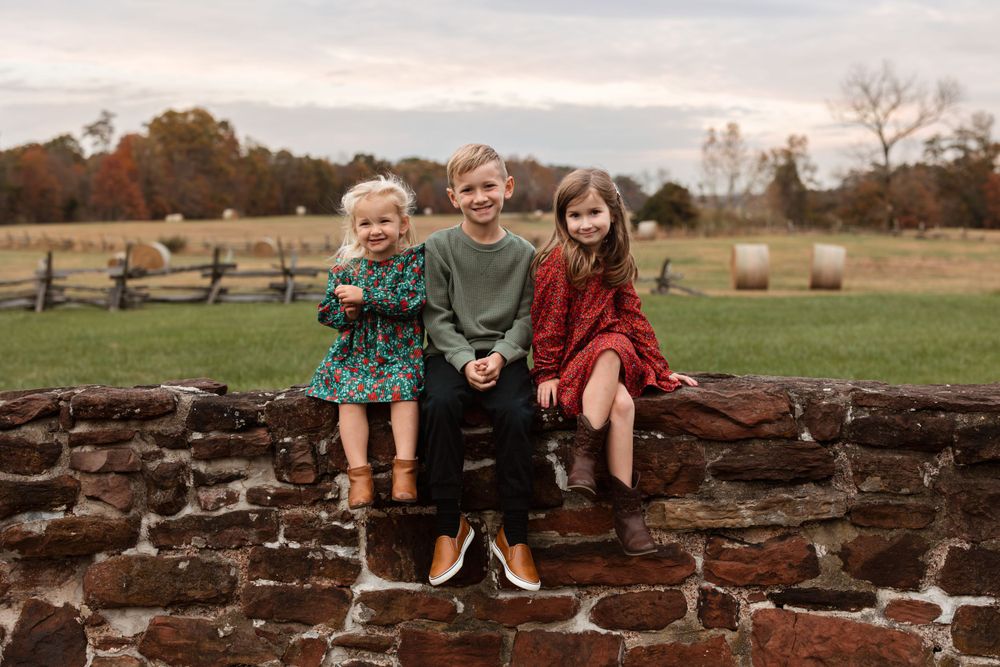 Three siblings sit together on stone wall with rustic countryside backdrop.