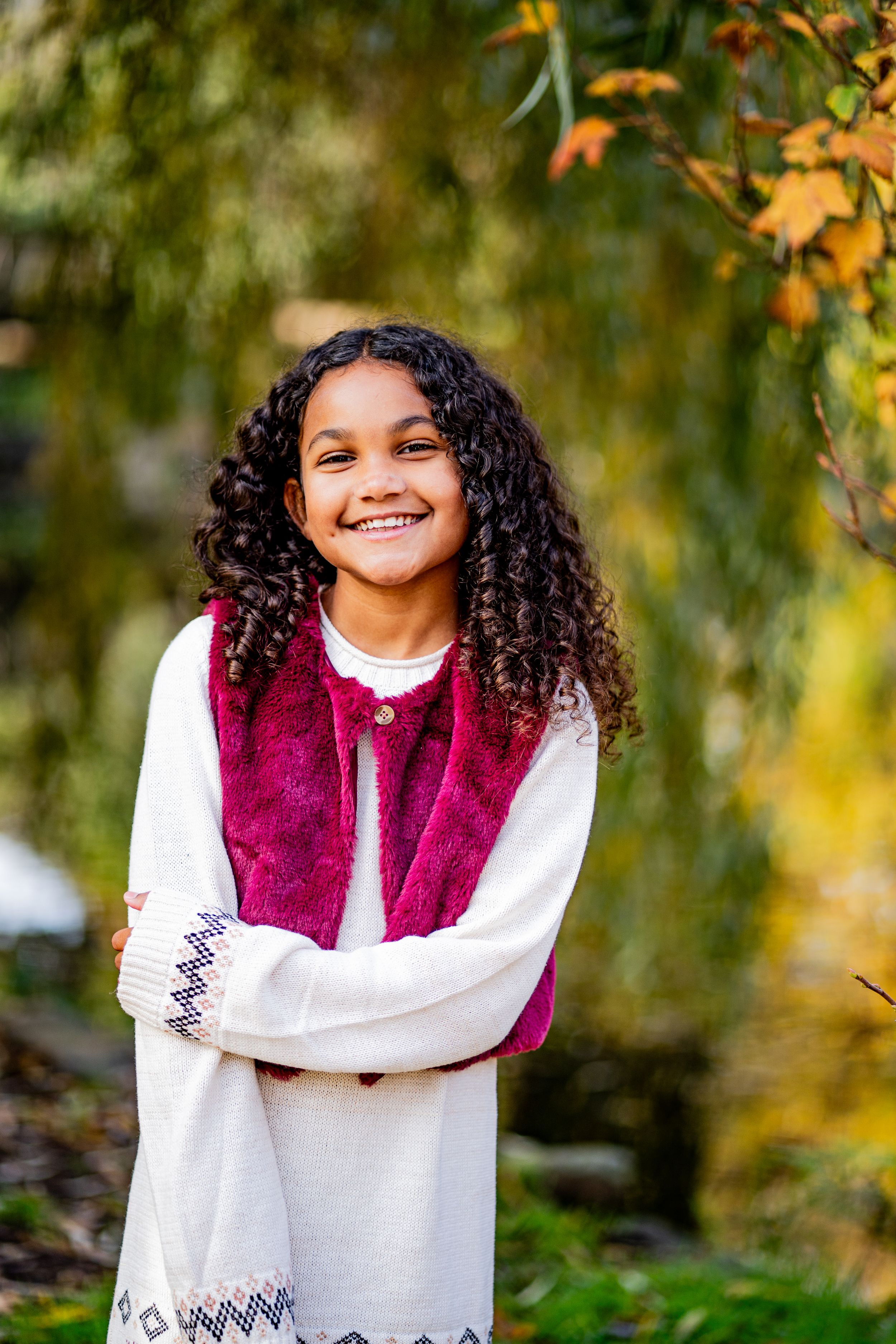 Person wearing a burgundy vest and white sweater poses outdoors among autumn foliage with natural lighting.