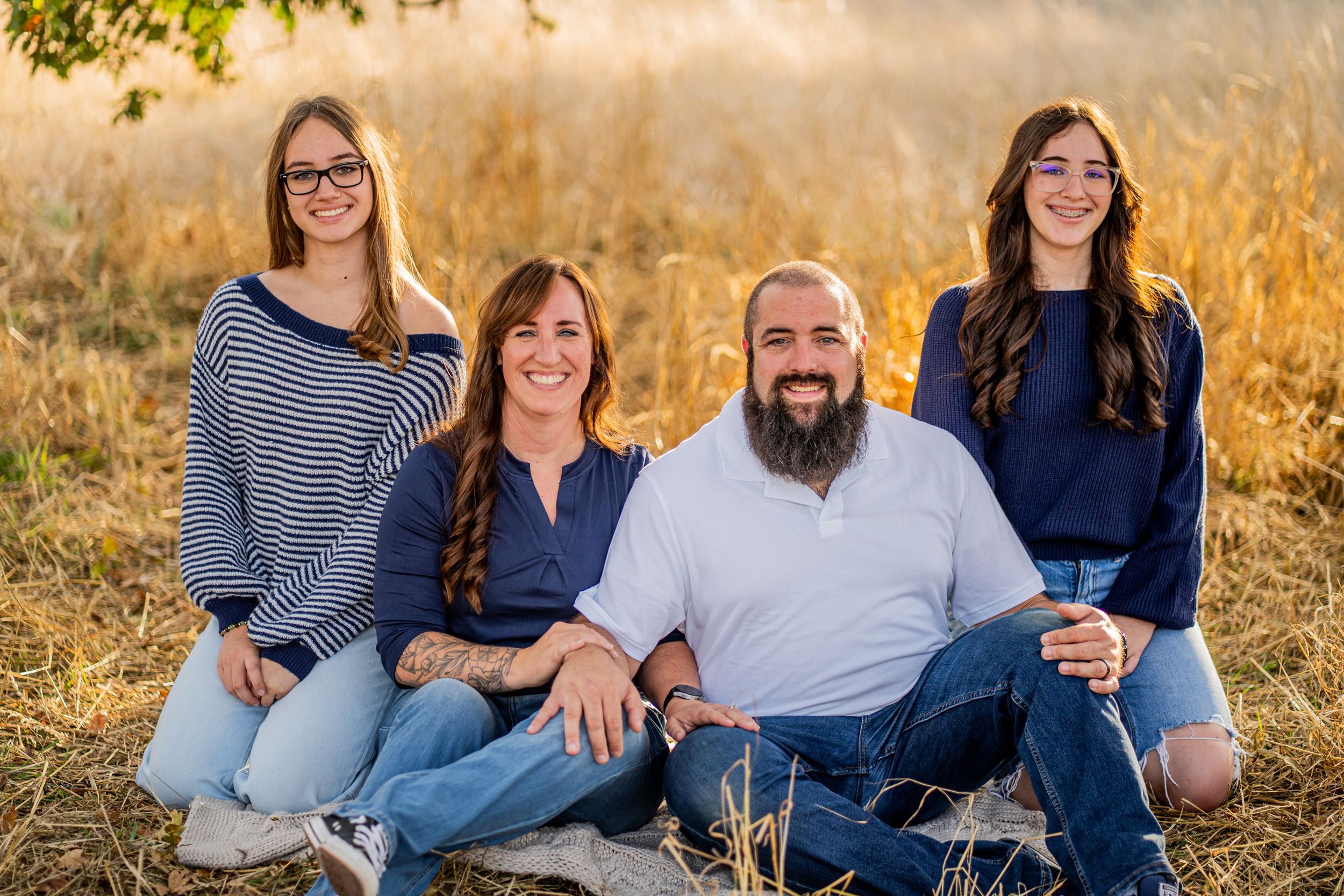 A family sits together in a golden autumn field during sunset wearing coordinated blue and white outfits.