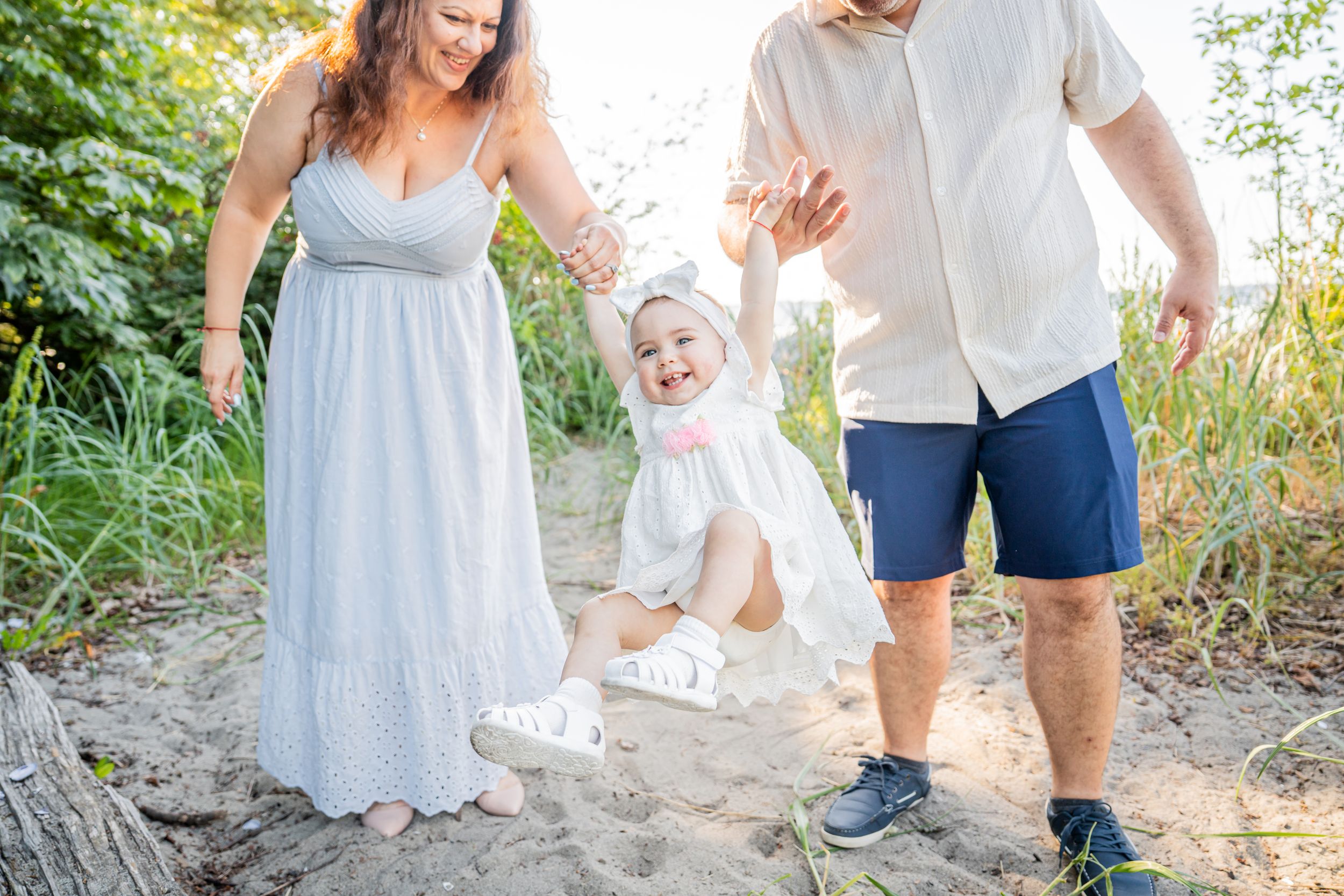 Family photo session with parents helping baby girl stand on sandy beach path surrounded by greenery.