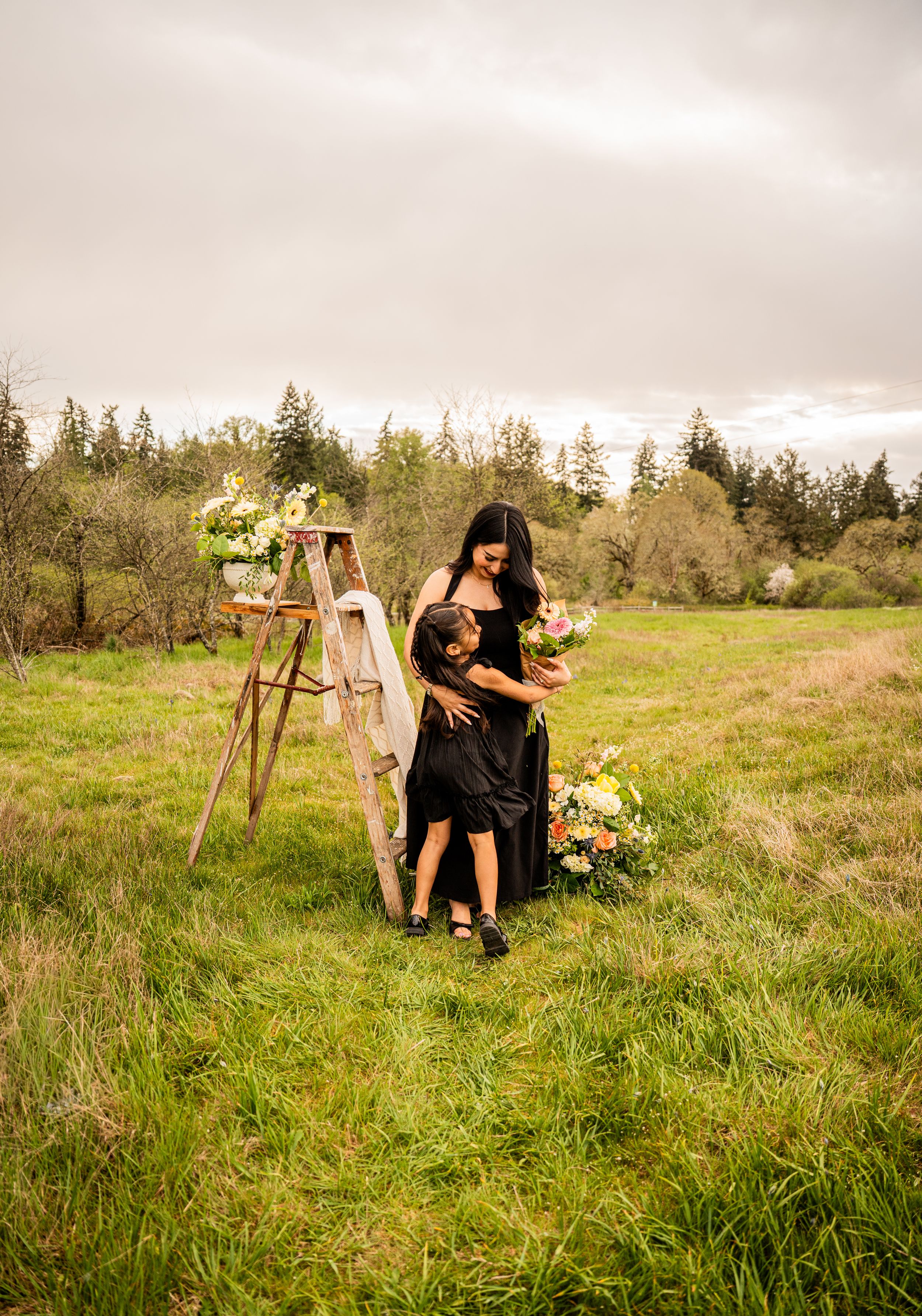 Woman in black dress hugging child in green meadow beside rustic ladder with floral arrangements and bouquet.
