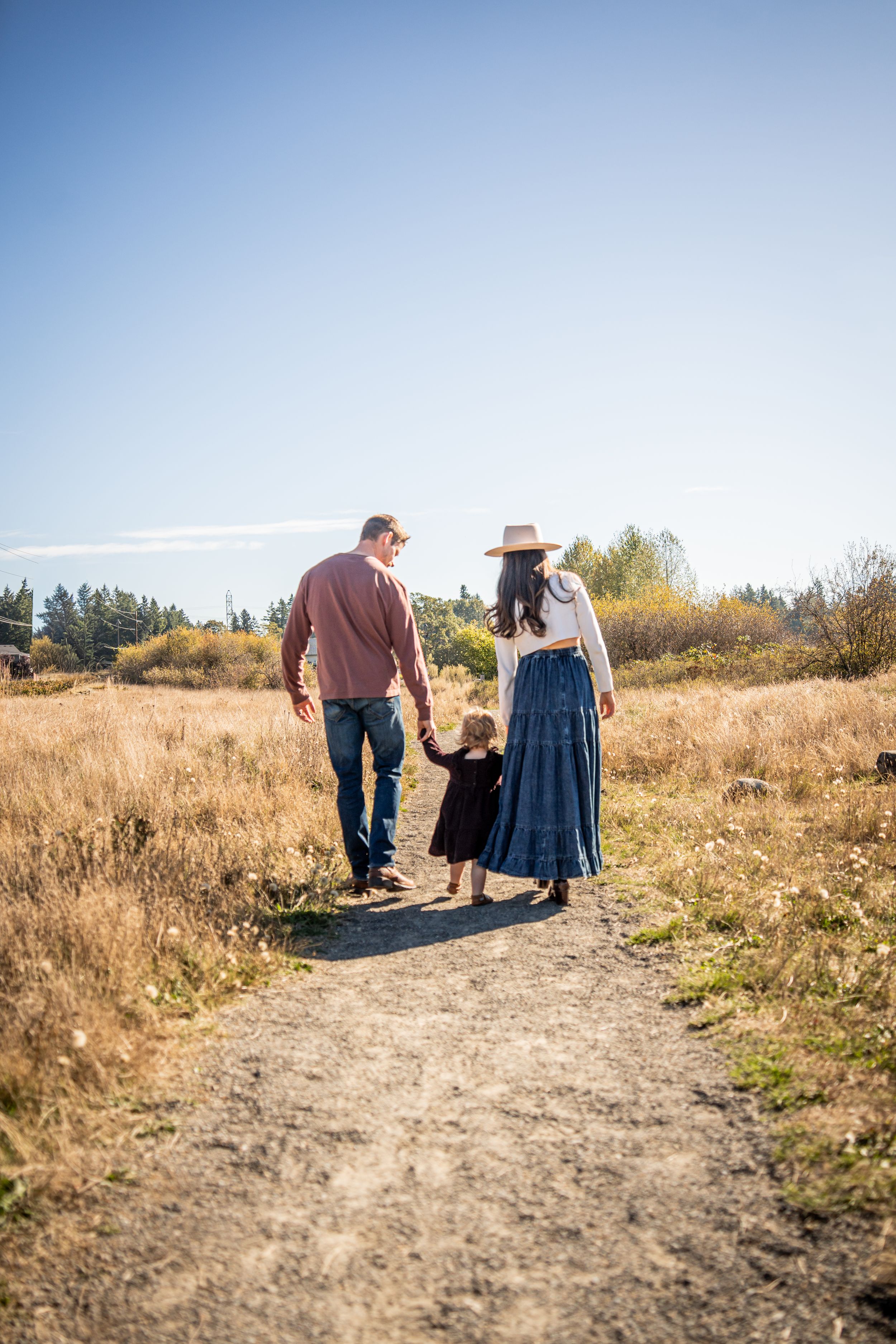 A couple walks together on a dirt path through golden autumn fields during sunset.