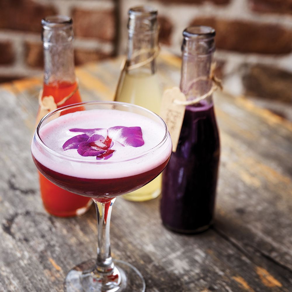 Purple-hued cocktail garnished with flower petals alongside bottles of colorful mixers on wooden surface.