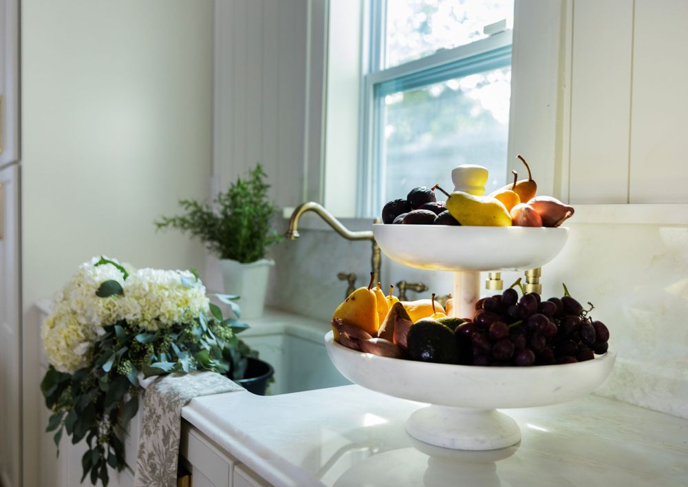Two-tiered white serving stand with fresh fruits and florals near bright window.