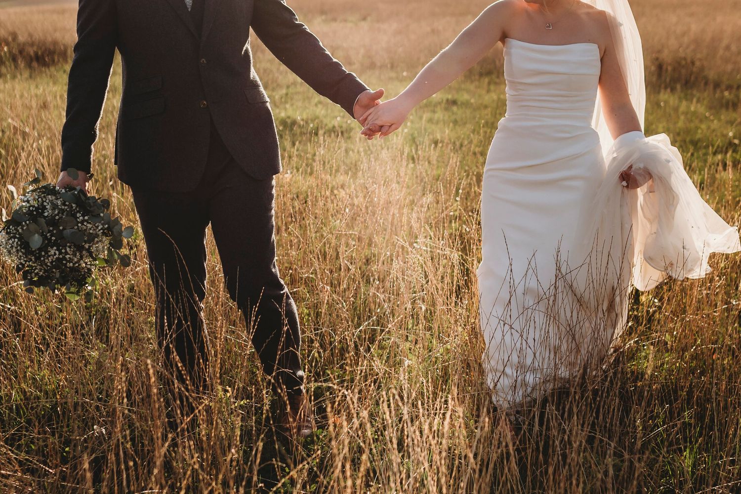 A silhouetted wedding couple holding hands while walking through a golden wheat field at sunset.
