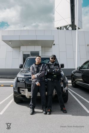 Two people posing in front of a black Land Rover Defender parked outside a modern white building.