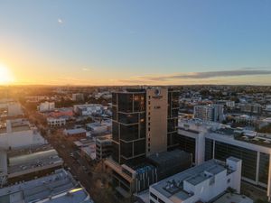 Aerial sunset view of a modern city skyline with a prominent glass office tower and urban sprawl under a clear blue sky.