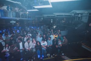 Panoramic view of a packed nightclub venue with blue atmospheric lighting and dancing crowd.