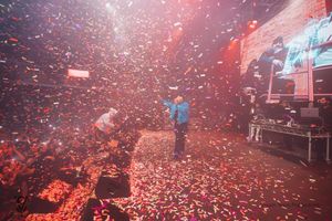 Concert crowd celebrates under red confetti and lights during an energetic live performance at an indoor arena.