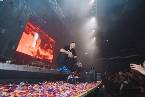 Concert performer jumps on stage with colorful confetti scattered below while large video screen glows red in background.