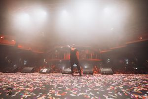 Silhouetted performer stands on stage surrounded by confetti in a smoky, red-lit concert venue.