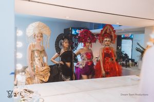 Performers in elaborate costumes and wigs pose together in front of a dressing room mirror with vanity lights.