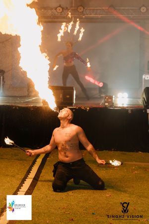 Fire performer kneeling on grass creates dramatic display with flaming props and red lighting effects.