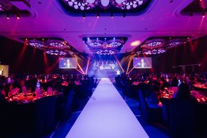 Elegant gala dinner venue with white runway, purple lighting, stage screens, and round tables in a grand ballroom.