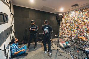 Musicians perform in a studio space with colorful mosaic-style wall art visible in the background.