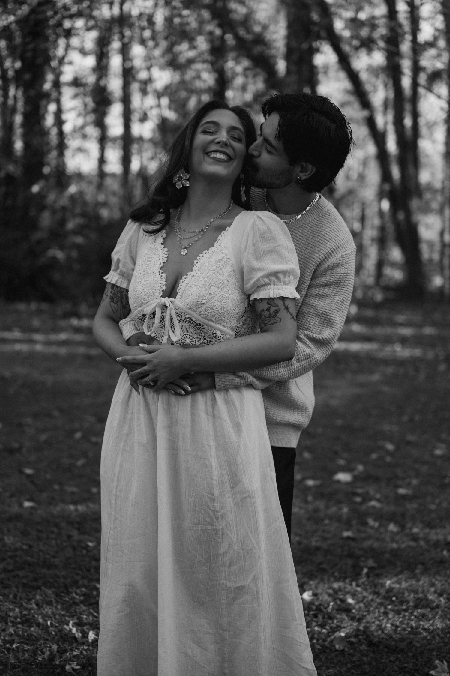 A joyful couple embraces in a wooded setting wearing vintage-inspired white dress and casual attire.