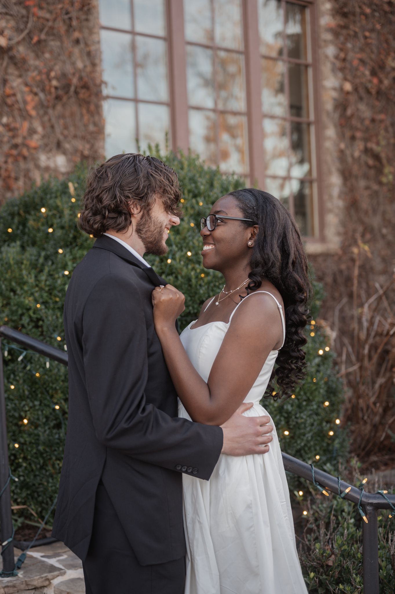 Couple sharing romantic moments against ivy-covered building with twinkling lights.