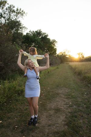 Sioux City, Iowa photographer. Joyful blonde mother carrying her toddler on her shoulders along a sunny nature trail at golden hour.