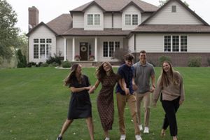 Sioux City, Iowa photographer. Happy family of five laughs and poses together on green lawn in front of large suburban home.