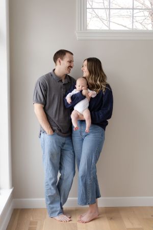 Sioux City, Iowa photographer. Young family with newborn baby poses barefoot by a bright window in a cozy, neutral-toned room.