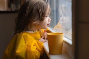Sioux City, Iowa photographer. Curious toddler in yellow raincoat gazes out a rainy window while holding a matching yellow cup.