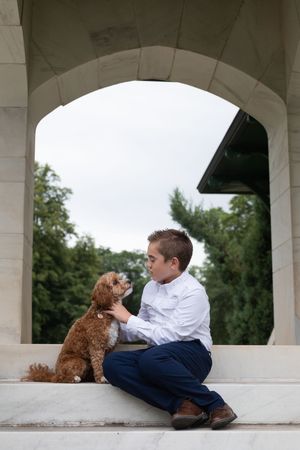 Sioux City, Iowa photographer. Young boy in white shirt and navy pants sitting with a fluffy brown cavapoo dog under a stone arch outdoors.